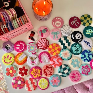 May include: A collection of colorful, round, hand-tufted coasters with various designs, including flowers, smiley faces, and geometric patterns. A lit candle and a pink storage crate filled with more coasters are also visible.