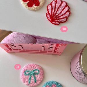 May include: A collection of colorful, embroidered coasters. Designs include a red flower on a cream background, a pink seashell, a pink bow, and a blue flower with a pink center. These decorative coasters are perfect for adding a touch of handmade charm to any home.