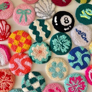 May include: A collection of colorful, round, hand-tufted coasters with various designs. The coasters feature patterns like a bow, a seashell, an eight ball, and a smiley face. The color palette includes pink, green, yellow, and blue, creating a vibrant and playful aesthetic.
