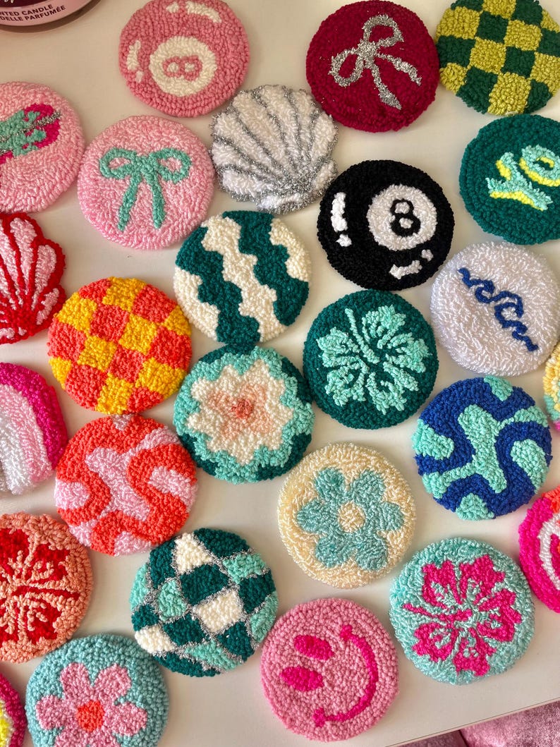May include: A collection of colorful, round, hand-tufted coasters with various designs. The coasters feature patterns like a bow, a seashell, an eight ball, and a smiley face. The color palette includes pink, green, yellow, and blue, creating a vibrant and playful aesthetic.
