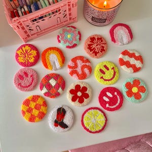 May include: A collection of colorful, handmade, circular coasters with various designs, including flowers, rainbows, and smiley faces. The coasters are arranged on a white surface, alongside a pink storage crate and a lit candle.