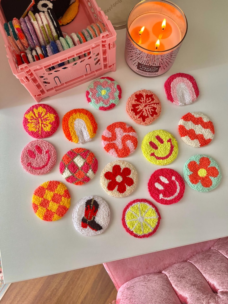 May include: A collection of colorful, handmade, circular coasters with various designs, including flowers, rainbows, and smiley faces. The coasters are arranged on a white surface, alongside a pink storage crate and a lit candle.