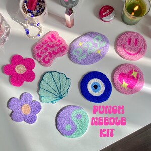 May include: A punch needle kit featuring a variety of colourful designs, including a flower, a seashell, an evil eye, a yin yang symbol, a smiley face, a heart, and a "Say It, Don't Say It Club" design. The kit is displayed on a white surface with a candle and other items.