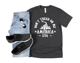 May include: A dark grey t-shirt with a white graphic of a snake with the text "Don't Tread On Me", a star, the word "America", and the year "1776".