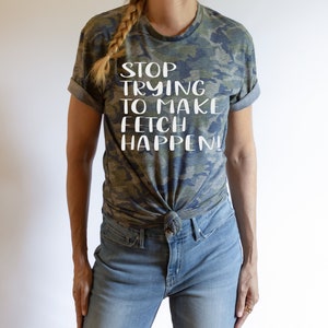 Könnte beinhalten: Eine Frau trägt ein T-Shirt mit Tarnmuster und dem Text "STOP TRYING TO MAKE FETCH HAPPEN!" in weißen Buchstaben. Sie trägt eine blaue Jeans und hat ihre Haare zu einem Zopf geflochten.