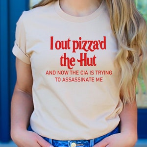Op de afbeelding: Een beige T-shirt met rode tekst die "I out pizza'd the Hut and now the CIA is trying to assassinate me" zegt.
