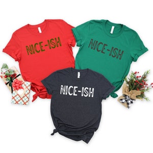 May include: Three t-shirts in red, green, and charcoal grey, each with the text "NICE-ISH" printed across the front in contrasting colours. The red shirt has green text, the green shirt has brown text, and the charcoal grey shirt has white text. Christmas-themed apparel.