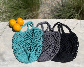 May include: Three crocheted mesh bags in teal, gray, and black. The bags are lying on a concrete surface with three oranges in the background.