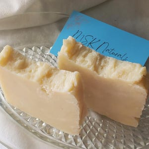 May include: Two bars of light beige handmade soap with textured tops, presented on a clear, decorative glass plate. A blue card with the text "MSK Naturals" is placed behind the soap.