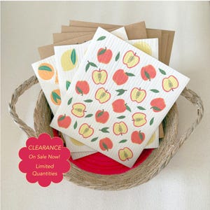 May include: A stack of square, white, textured cellulose dishcloths featuring a repeating pattern of red and yellow apples with green leaves, resting in a woven basket. A pink scalloped sign displays the text CLEARANCE On Sale Now! Limited Quantities.