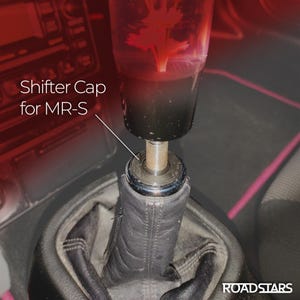 Peut inclure: Gros plan sur le levier de vitesse d'une voiture avec un pommeau personnalisé. Le pommeau est transparent avec un motif rouge à l'intérieur. Le texte "Shifter Cap for MR-S" est visible en blanc. Le soufflet est en cuir noir.