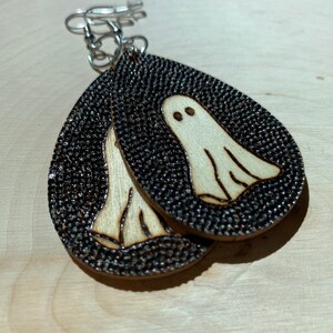 May include: A pair of wooden earrings with a black and white ghost design. The earrings are shaped like teardrops and have silver hooks.