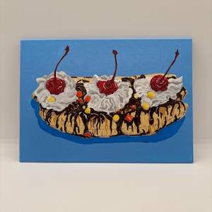 May include: A painting of a banana split on a blue background. The dessert features a split banana base, topped with whipped cream, cherries, chocolate sauce, and colorful sprinkles. The artwork is on a rectangular canvas.