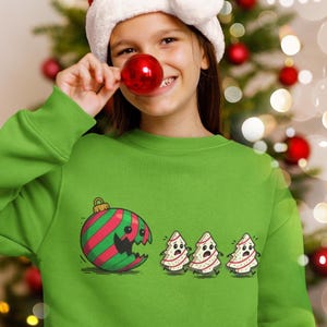 May include: Green sweatshirt with a Christmas ornament and Christmas tree graphic. The ornament is red and green striped with a broken section. Three cartoon Christmas trees are also on the sweatshirt. A person is wearing a Santa hat and holding a red ornament.