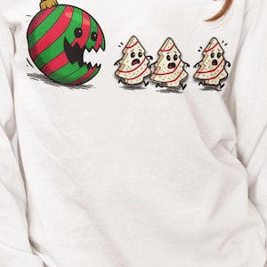 May include: White long-sleeve shirt featuring a cartoon design. The design includes a red and green striped ornament with a broken section and three frightened Christmas tree cookies. The ornament has a gold hook.