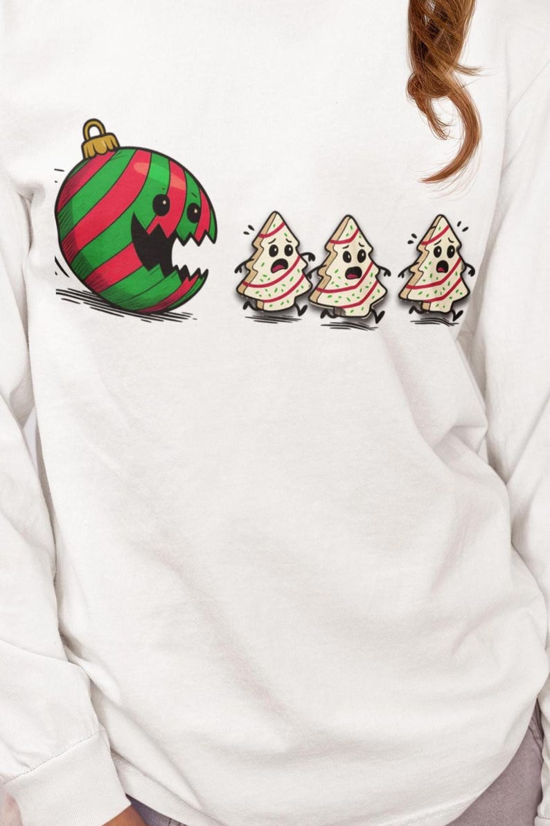 May include: White long-sleeve shirt featuring a cartoon design. The design includes a red and green striped ornament with a broken section and three frightened Christmas tree cookies. The ornament has a gold hook.