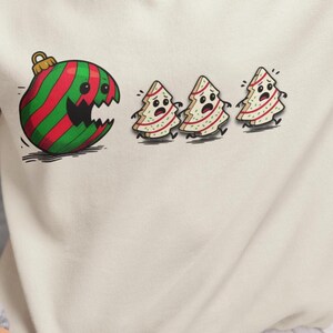 May include: Off-white sweatshirt featuring a cartoon graphic. The graphic depicts a large, green and red striped Christmas ornament with a bite taken out of it, chasing three frightened Christmas trees. The design has a whimsical, holiday theme.