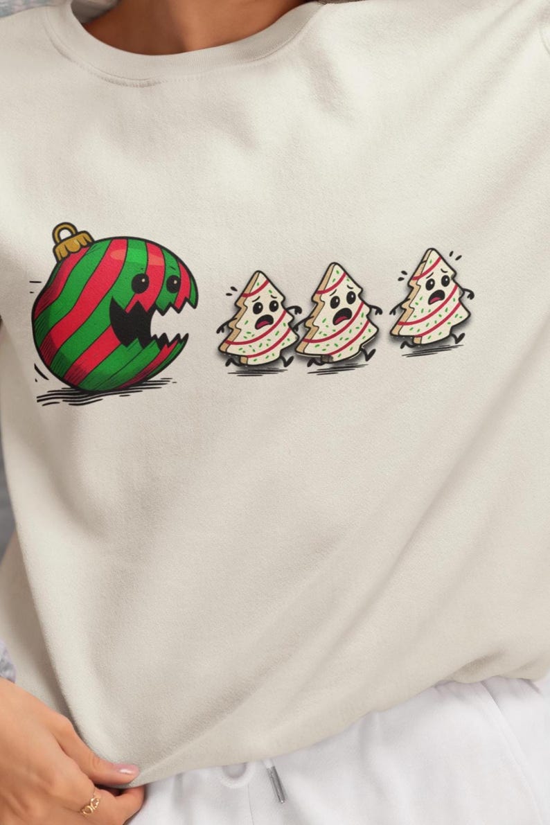 May include: Off-white sweatshirt featuring a cartoon graphic. The graphic depicts a large, green and red striped Christmas ornament with a bite taken out of it, chasing three frightened Christmas trees. The design has a whimsical, holiday theme.