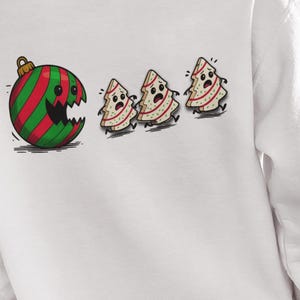 May include: White sweatshirt featuring a cartoon design. A large Christmas ornament with red and green stripes has a gaping mouth, chasing three frightened Christmas tree figures. The design is on the back of the sweatshirt.