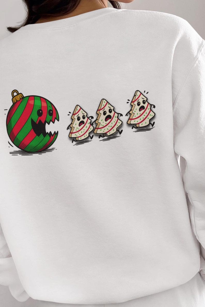 May include: White sweatshirt featuring a cartoon design. A large Christmas ornament with red and green stripes has a gaping mouth, chasing three frightened Christmas tree figures. The design is on the back of the sweatshirt.
