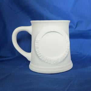 May include: A white ceramic mug with a large handle and a circular embossed design. The design features the words "IN DEFENSE OF FREEDOM" around the edge. The mug is set against a blue background.