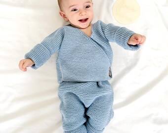Op de afbeelding: Een baby draagt een lichtblauwe gebreide wikkelvest en bijpassende broek. De outfit heeft een textuur breipatroon en een knoopsluiting. De baby ligt op een witte ondergrond en lacht. Een houten schijf met tekst is ook aanwezig.