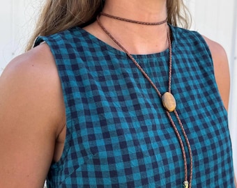 Puede incluir: Una corbata bolo con un cordón de cuero trenzado marrón, un colgante central ovalado de piedra ojo de tigre y dos puntas metálicas doradas. El accesorio se lleva sobre una prenda sin mangas con un patrón de cuadros vichy en azul cerceta y negro.