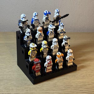 Może przedstawiać: Kolekcja figurek Lego Star Wars, w tym różnych żołnierzy-klonów, prezentowana na czarnym, schodkowym stojaku. Figurki mają białe zbroje z niebieskimi, pomarańczowymi, żółtymi i czerwonymi akcentami. Niektóre figurki trzymają blastery.