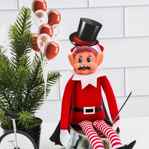 Peut inclure: Un lutin en peluche rouge et blanc portant un chapeau haut de forme noir avec une bande rouge, assis sur un seau argenté. Le lutin a une barbe blanche, une moustache noire et des yeux bleus. Le lutin tient une baguette noire. Il y a des ballons rouges et blancs en arrière-plan.