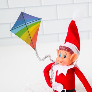 May include: A red and white elf doll with a white hat and a red suit is holding a rainbow kite with a silver string.