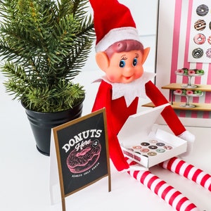 May include: A Christmas elf doll wearing a red and white striped outfit is holding a box of donuts. The box has a pink and white striped design and says "Donut Shop". The elf is sitting in front of a small chalkboard sign that says "Donuts Here $2 Always Only Fresh" with a pink and white donut graphic.