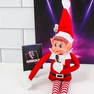 May include: A red and white elf doll holding a microphone in front of a black and purple background with spotlights. The elf is wearing a Santa hat and has a white beard. The background has the text "Karaoke Party" on a black sign.