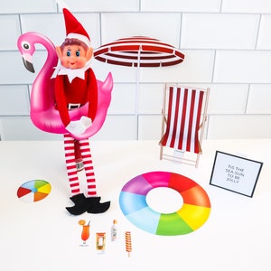 May include: A Christmas elf doll wearing a red and white striped outfit and a Santa hat is sitting on a pink inflatable flamingo. The elf is holding onto the flamingo with both hands. There is a red and white striped beach chair, a rainbow inflatable ring, a beach ball, a sign that says "Tis the sea, sun to be jolly", and a few other beach-themed accessories in the image.