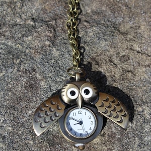 May include: A gold owl pocket watch with a white face and black hands. The watch is on a gold chain and has a detailed owl design.