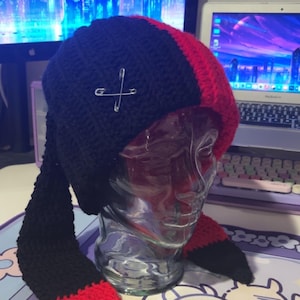 May include: A black and red crocheted hat with long bunny ears. The hat has two safety pins crossed over each other on the top.