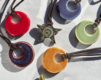 May include: A collection of seven ceramic pendants on dark brown leather cords. Five pendants are circular with a central hole in red, blue, light green, dark purple, and orange. Two star-shaped pendants feature a spiral design in blue and lime green.
