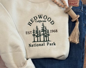 May include: A beige sweatshirt with a green embroidered design that says "Redwood California Est 1968 National Park".