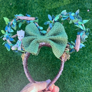 May include: Green and brown fabric headband with a green bow and a small figurine of a character from the movie Guardians of the Galaxy. The headband is decorated with green leaves and brown twigs.