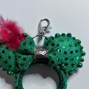 以下が含まれることがあります： 緑色のスパンコールのミニーマウスの耳に、赤い羽根が飾られています。耳には、バッグやキーホルダーに取り付けられるシルバーの金属製の留め具が付いています。