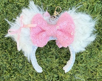 May include: A white faux fur keychain featuring a pink sequined lace bow and a small pink ribbon tie. The accessory includes a silver-toned metal lobster clasp attached to the top, with a white sequined trim along the bottom edge of the fur.
