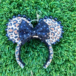 以下が含まれることがあります： ヒョウ柄のミニーマウスの耳のヘアバンド。黒いスパンコールのリボンと黒いビーズトリムが付いています。耳は丸く、ヘアバンドは黒です。シルバーのキーホルダーが付いています。