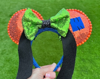 Op de afbeelding: Een paar oranje en groene, met pailletten bedekte Minnie Mouse oren met een zwarte strik en een blauwe hoofdband. De oren hebben een blauwe patch aan de ene kant.