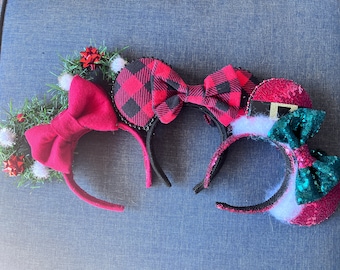 May include: Three festive headbands with ear shapes. One features a red bow and greenery, another has a red and black plaid bow, and the third is designed as a Santa hat with a green sequined bow. All have black headbands.