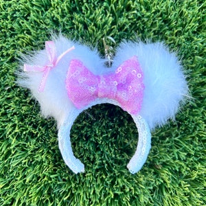 以下が含まれることがあります： ピンクのキラキラしたリボンが付いた白いふわふわのミッキーマウスの耳のヘッドバンド。