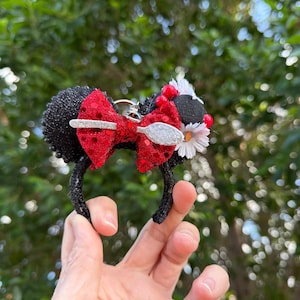 Puede incluir: Una diadema negra con dos orejas circulares cubiertas de purpurina negra. Un lazo rojo de lentejuelas se sitúa entre las orejas, decorado con un dije de cuchara de purpurina plateada, pequeñas cuentas rojas y flores de margarita blancas.