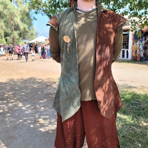 May include: A person wearing a Renaissance-style outfit. The outfit includes a green and brown suede vest, a textured olive green shirt, and patterned rust-colored pants. The outfit is accessorized with a necklace and a small wooden pin.