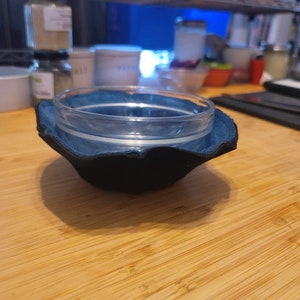 May include: A clear glass bowl sits inside a dark blue and black fabric bowl cozy. The cozy has a scalloped edge and is resting on a light brown wooden surface. The background is blurred, showing kitchen items.