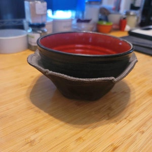 May include: A black and red ceramic bowl sits inside a brown, leather-like bowl cozy. The bowl cozy has a unique, folded design. The bowl is on a wooden surface, with other kitchen items in the background.