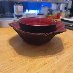 May include: A black bowl with a red interior sits inside a burgundy fabric bowl cozy. The cozy has a square shape with folded edges. The bowl and cozy are on a light-colored wooden surface, creating a contrast of colors and textures.
