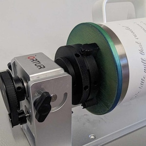 May include: A white tumbler with a green and silver lid is mounted on a rotating platform. The tumbler has a handwritten inscription that reads "Our little mill that could..." and includes names and dates. The platform is attached to a silver base with a black knob and the word "Ortur" printed on the side.